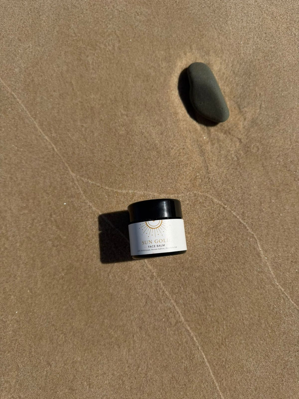 Sun Gold Face Balm jar on wet beach sand beside a smooth stone