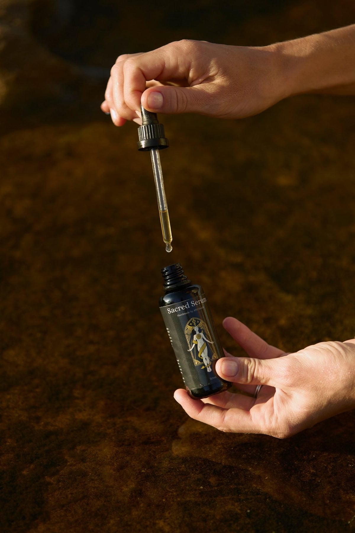 Sacred Serum dropper of ozonated macadamia oil held over a coastal rock pool