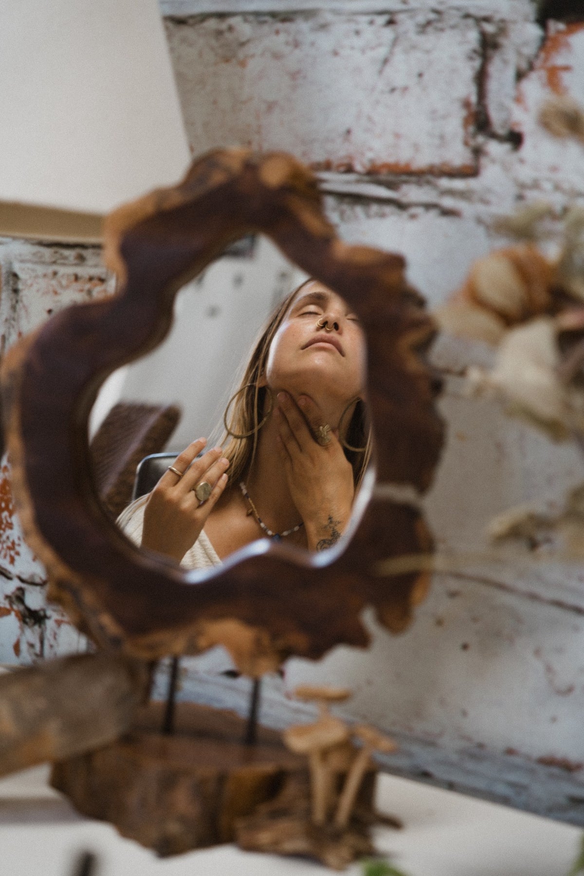 Eyes closed, hands resting at the throat, reflected in a raw timber mirror