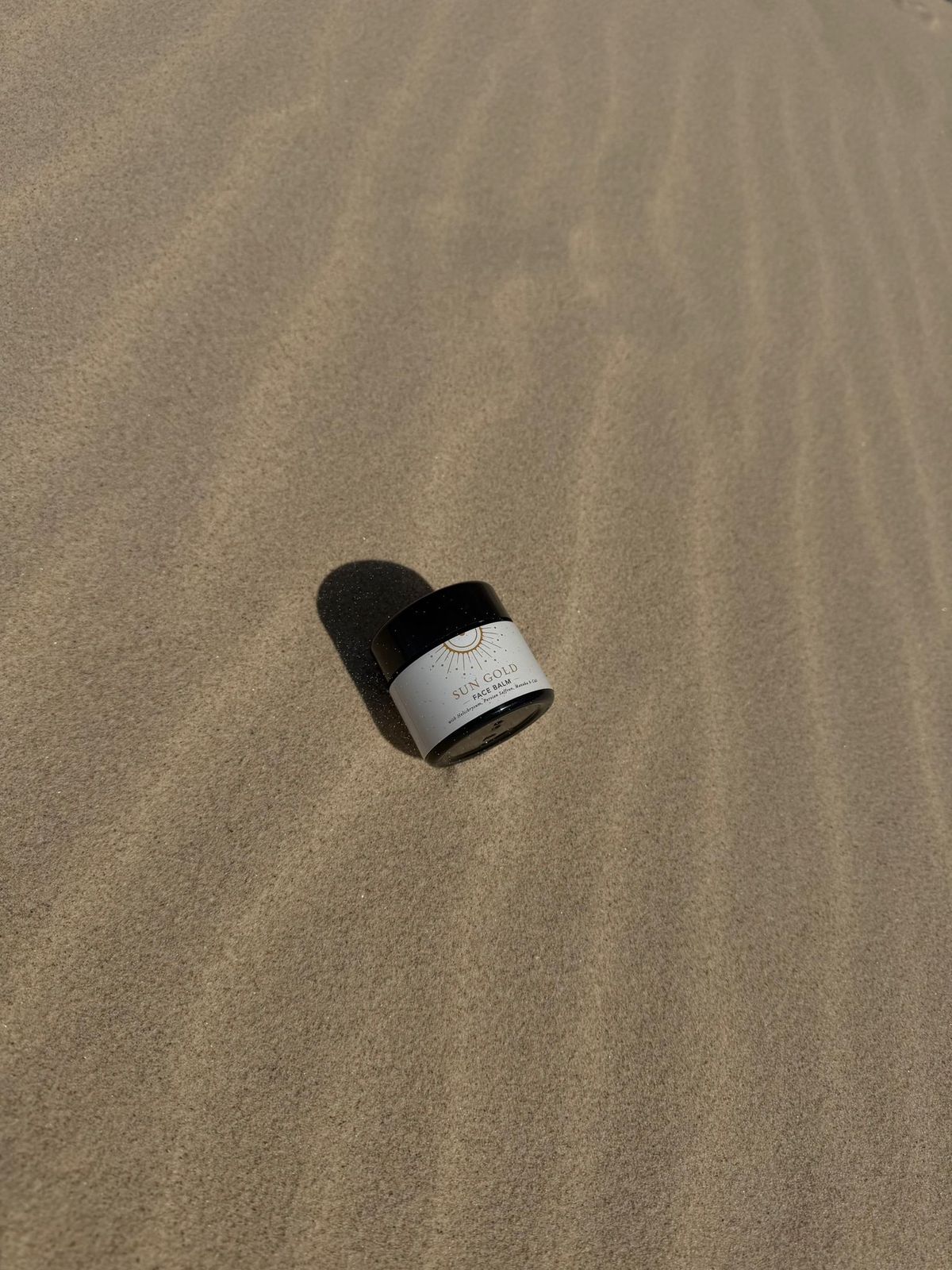 A jar of Sun Gold Face Balm alone on wide, wind-rippled beach sand